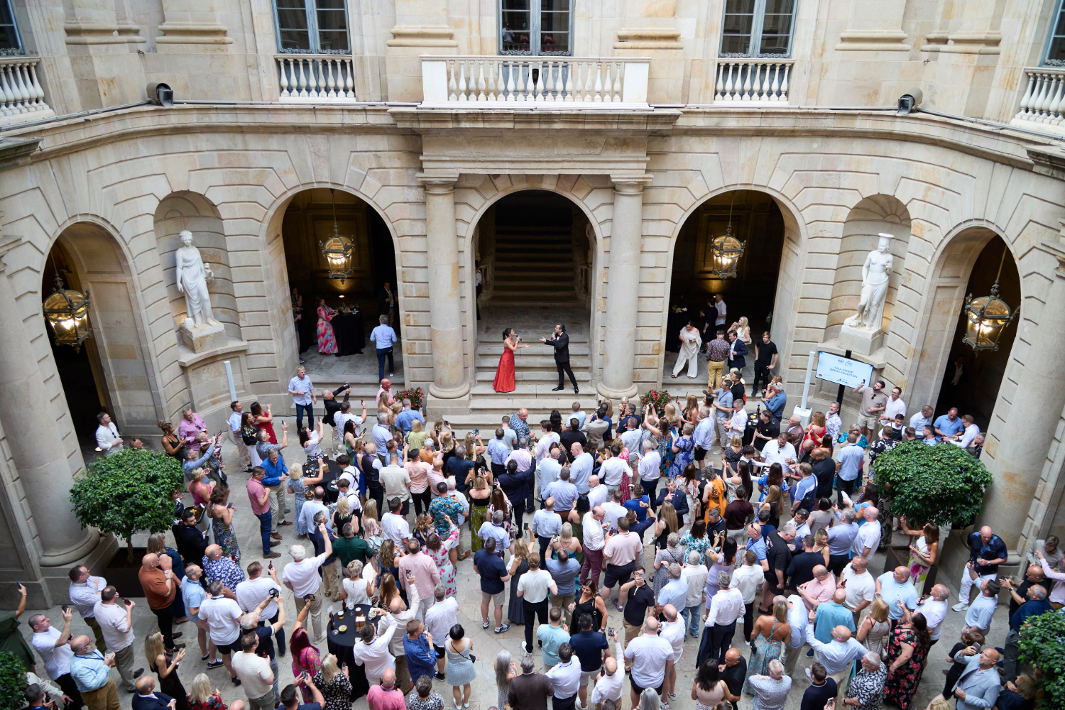 A large crowd gathers in the courtyard of a grand stone building with arches, statues, and balconies, watching two people, one in a red dress and one in black, performing near the entrance.