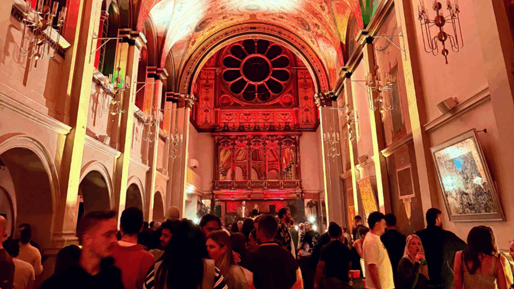 A crowd of people socializes and mingles inside a dimly lit, ornate hall with high arched ceilings and chandeliers, as colorful lights illuminate the space, creating a lively, festive atmosphere perfect for a private luxury event.