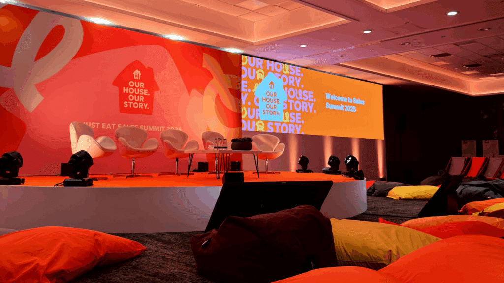 A brightly lit conference stage with four white chairs, a small table, and a large orange backdrop reading Our House. Our Story. Colorful bean bags offer relaxed seating at this private luxury event. The screen displays Welcome to Sales Summit 2025.