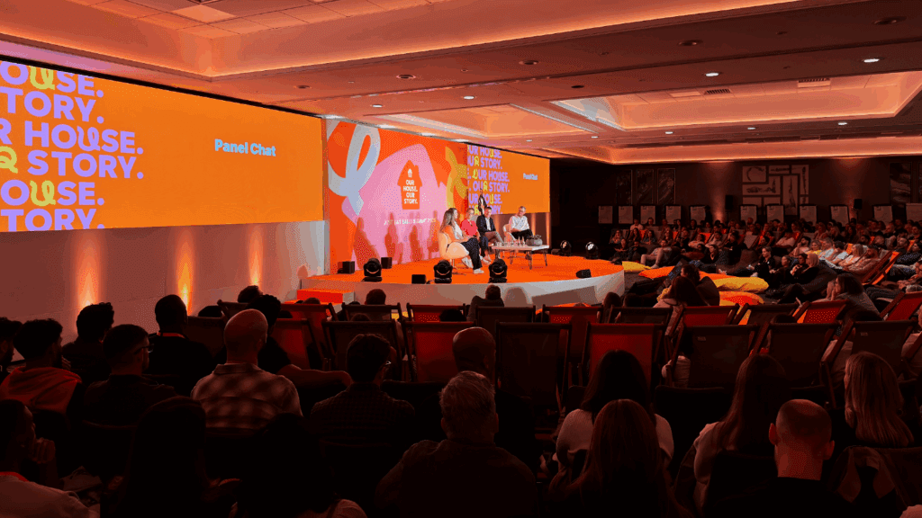 A large audience watches a panel discussion on a brightly lit stage with orange decor and “YOUR HOUSE. YOUR STORY.” displayed on screens at a private luxury event.