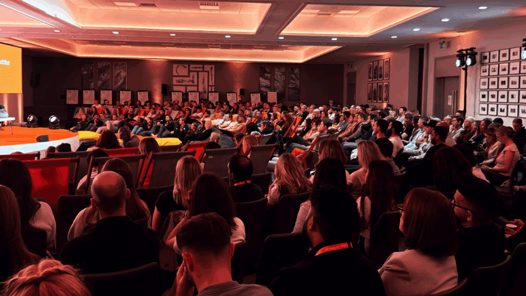 A large audience sits in rows of chairs, attentively watching a presentation on stage at a private luxury event in a brightly lit conference room with orange lighting and framed pictures on the walls.