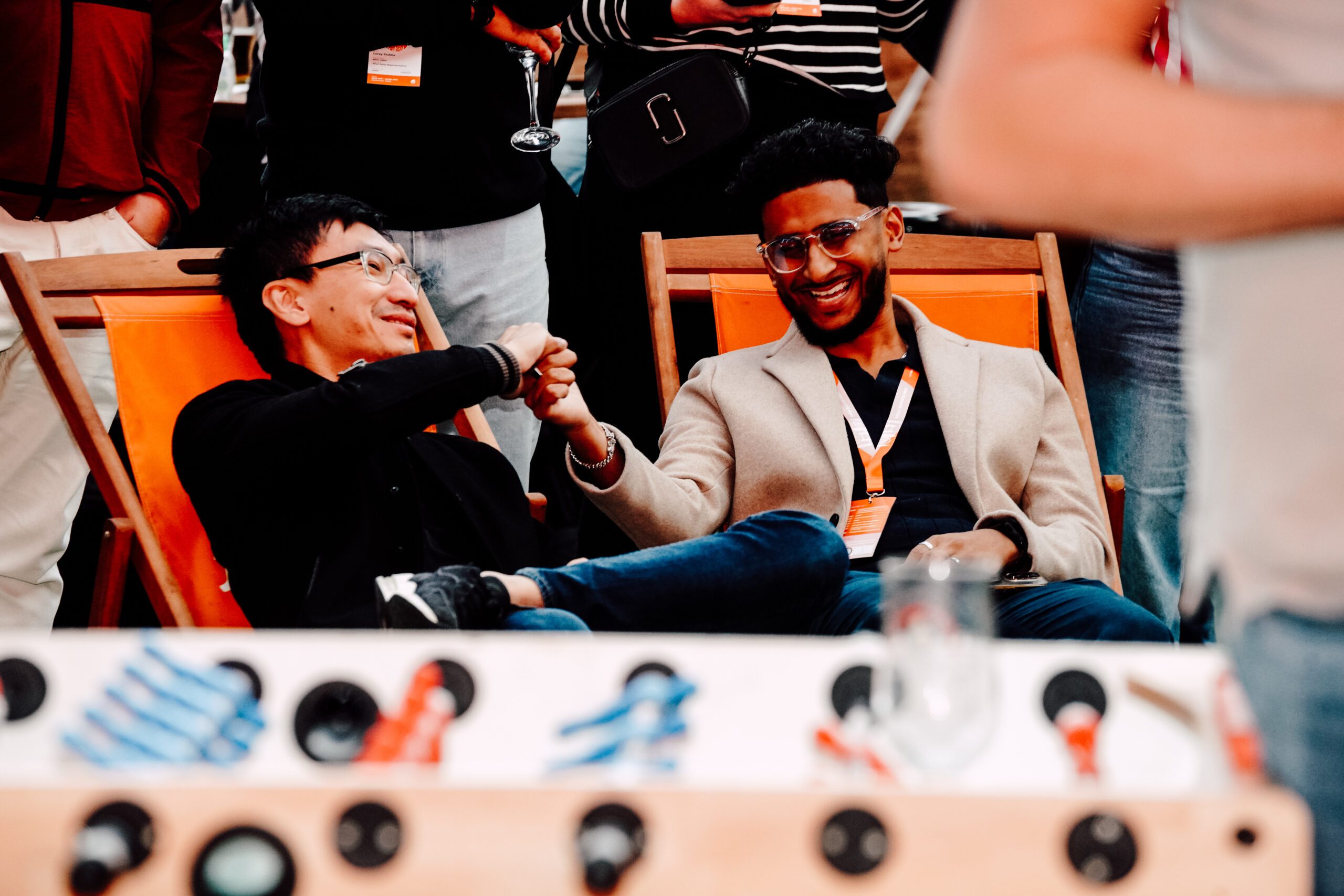 Two men sitting on orange lounge chairs smiling and fist-bumping, wearing conference badges. People stand in the background, and the foreground is blurred with part of a foosball table and empty glass visible.