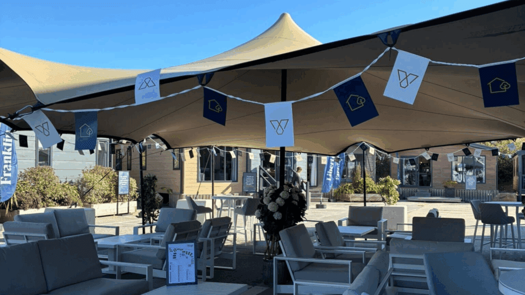 An outdoor seating area with white tables and chairs under large beige canopies, perfect for a product showcase. Decorative banners hang across the space, with modern buildings and blue sky in the background.