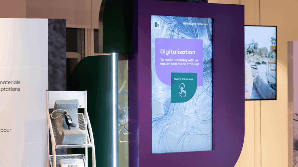 A large interactive digital display with the word Digitalisation and a touch symbol stands in a modern exhibition space at the BSL Conference, next to a shelf holding bricks and a plugged-in device.