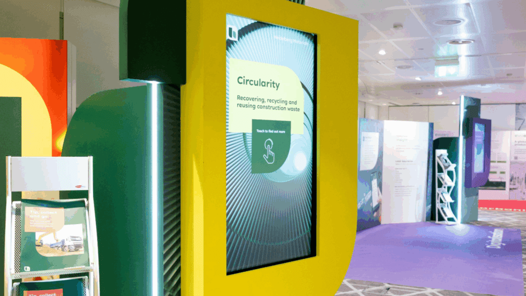 A digital display in a bright yellow frame shows information about circularity at the BSL Conference, focusing on recovering, recycling, and reusing construction waste at an indoor exhibition with informational panels and brochures.