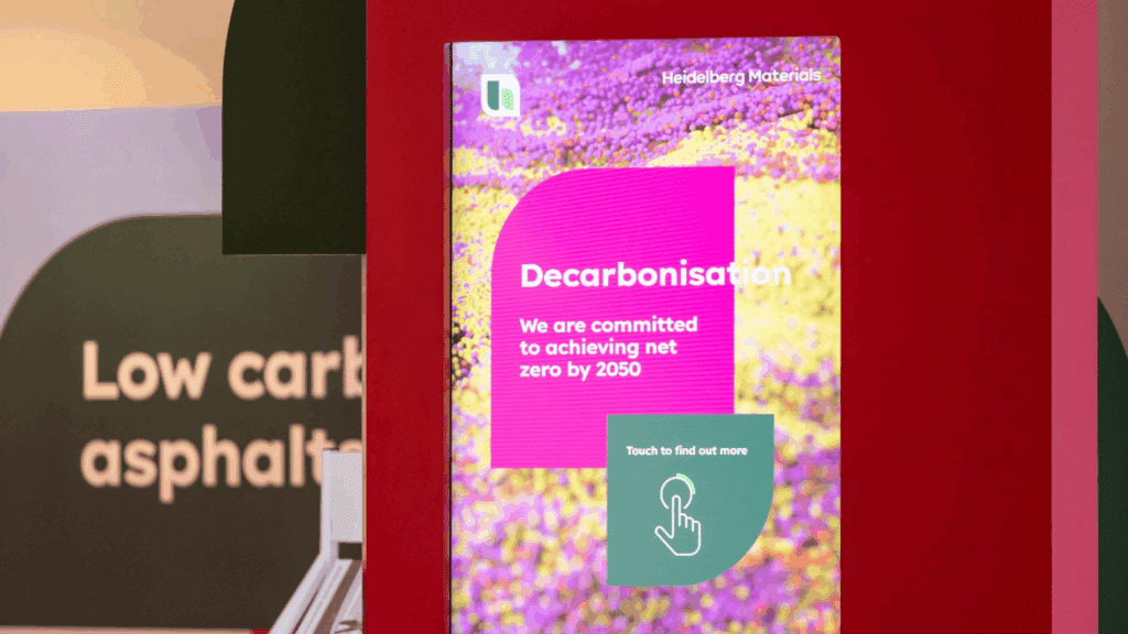 A digital display at the BSL Conference reads Decarbonisation: We are committed to achieving net zero by 2050 with a button that says, Touch to find out more. Colorful abstract background, part of a Low carbon asphalt booth.