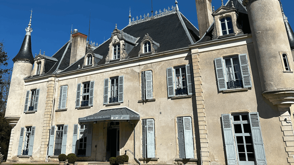 A large, elegant French chateau with cream-colored walls, blue shutters, tall arched windows, and pointed turrets hosts a Sustainability Conference beneath intricate rooftop detailing and a clear blue sky.