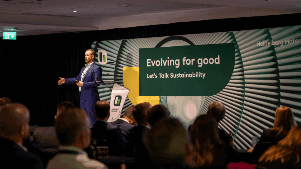 A man in a suit speaks on stage at the BSL Conference with a banner behind him that reads, Evolving for good - Let's Talk Sustainability. The audience is seated and listening attentively.
