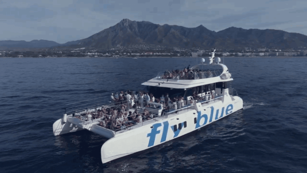 A large catamaran with “fly blue” written on the side sails on calm blue water, crowded with people enjoying a Sustainability Conference. In the background, a coastal city and mountains are visible under a clear sky.