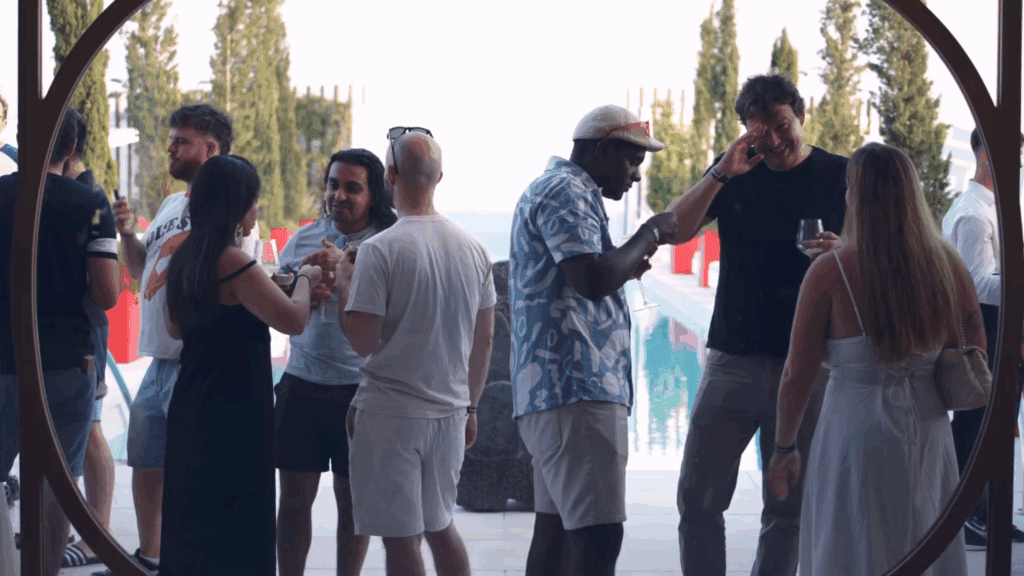 A group of people stand and talk near a pool outdoors at a Sustainability Conference, some smiling and laughing in casual summer clothes, with trees in the background.