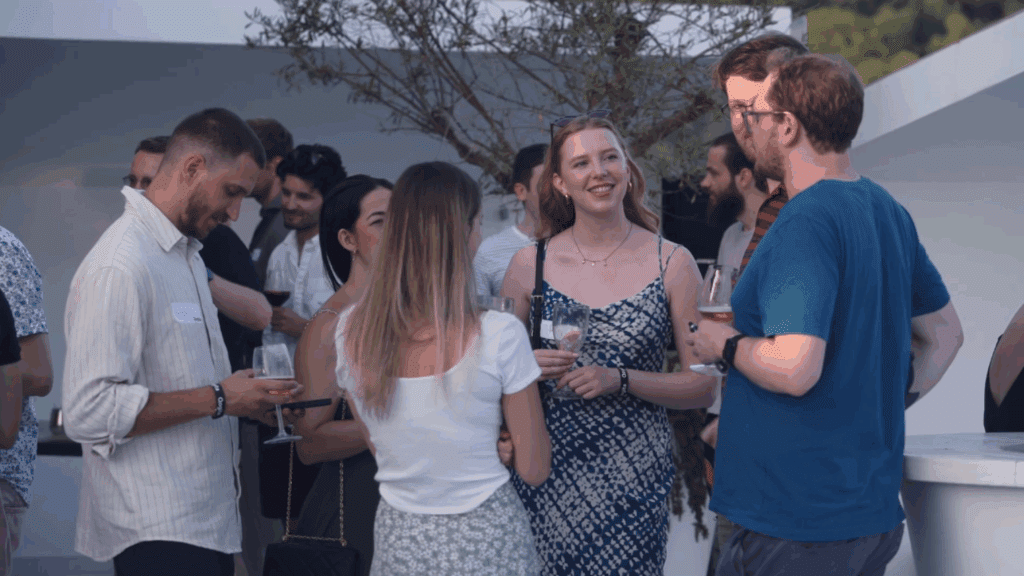 A group of people are gathered outdoors at a Sustainability Conference, socializing and holding drinks. They are casually dressed and appear to be enjoying a friendly conversation on a sunny day, with trees and other guests in the background.