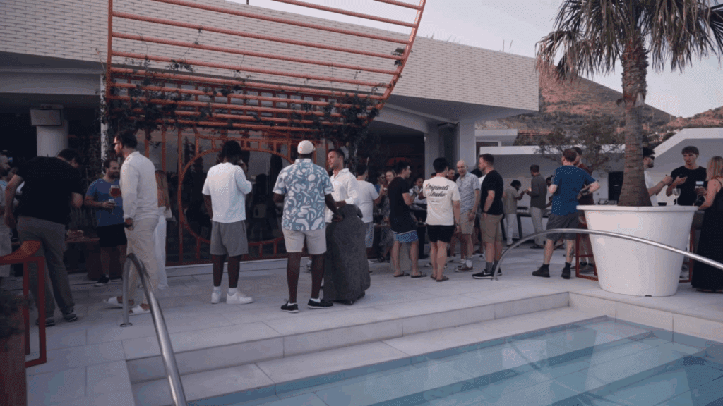 A group of people socializing by a modern outdoor pool area during a Sustainability Conference, some chatting near steps under a partial pergola with greenery, with palm trees and hills in the background.