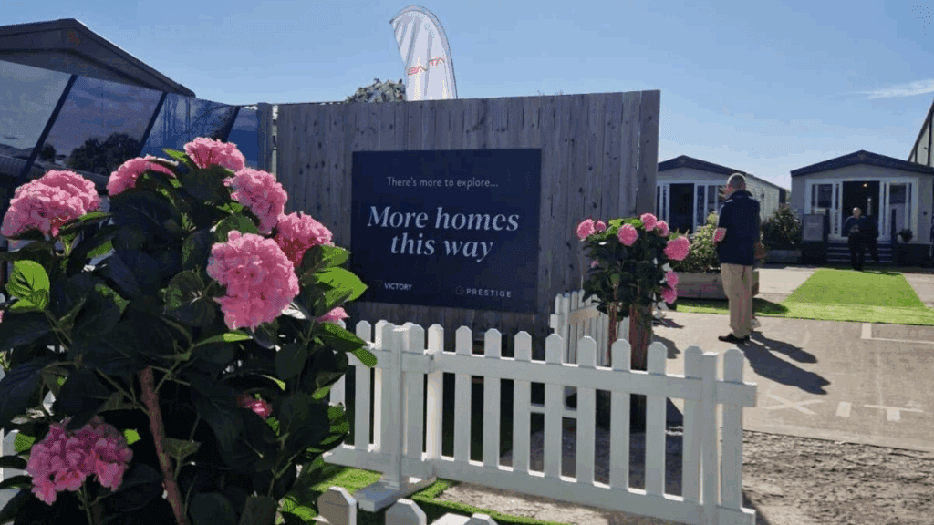 A sign reading “More homes this way” stands behind a white picket fence with blooming pink flowers, creating a charming product showcase. In the background, two people walk toward modern homes on a sunny day.