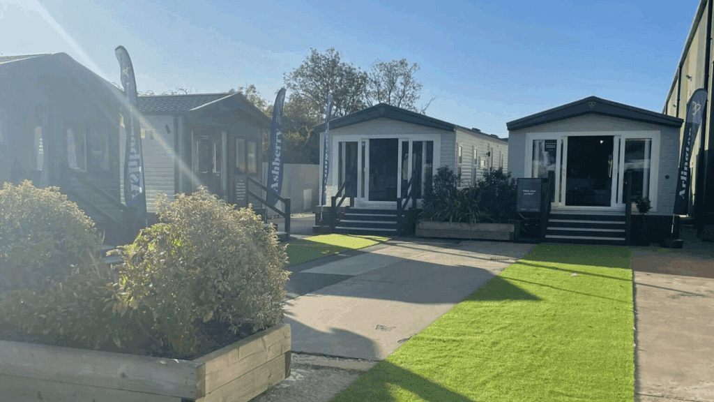 Three small, modern prefabricated homes are displayed on artificial grass with a clear blue sky above. Flags and plants in front highlight the area as a stylish product showcase or showroom for potential buyers.