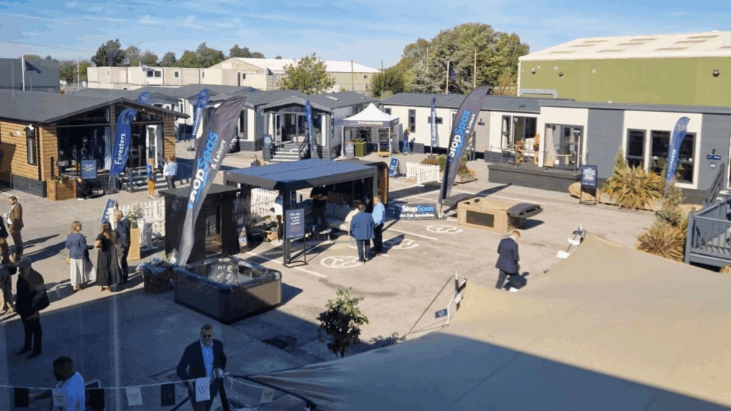 An outdoor product showcase at a modular building showroom, with people walking and talking among display cabins, flags, booths, and informational signs under a clear sky.