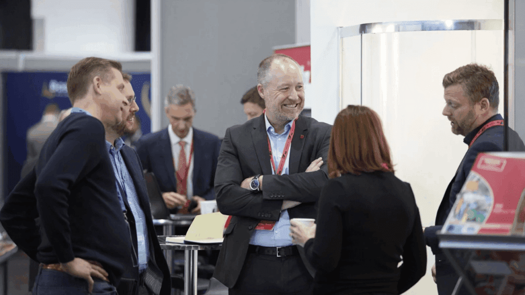 A group of professionally dressed people stand together in conversation at a business event or conference. One man in the center, smiling with his arms crossed, is surrounded by others near eye-catching exhibition stand design displays in the background.