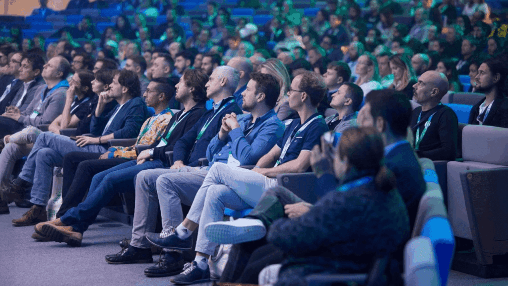 A large audience of people sits attentively in rows of seats at an indoor event, with blue lighting illuminating the scene, expertly arranged by the conference organiser.