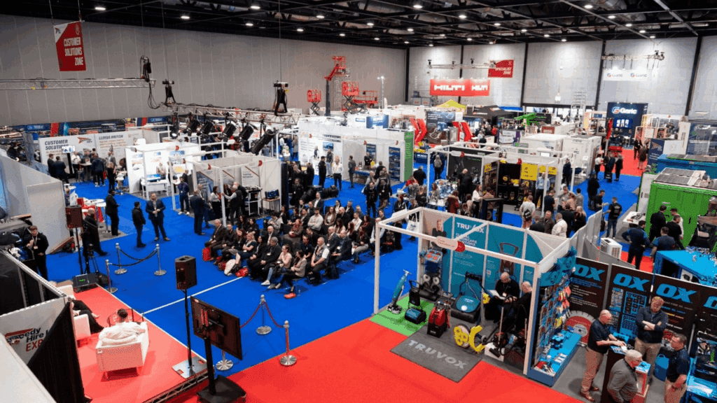 A busy trade show or convention featuring modern exhibition stand design, with numerous booths, product displays, and groups of people walking, talking, and seated in a large, well-lit indoor exhibition hall with blue carpeting.