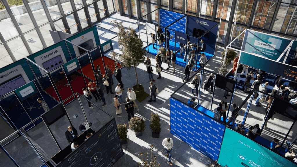 Aerial view of a modern indoor event with people walking and talking near innovative exhibition stand designs, blue and red displays, large windows, and sunlight streaming into the spacious hall.