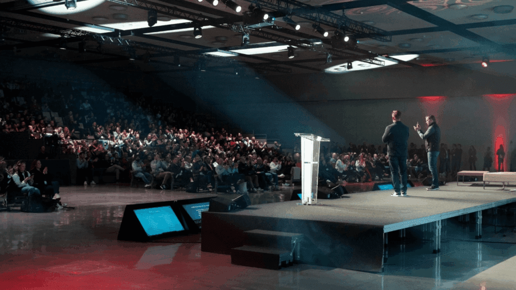 Two speakers, introduced by the conference organiser, stand on a stage in front of a large seated audience, with spotlights illuminating the stage and presentation screens visible on the floor.