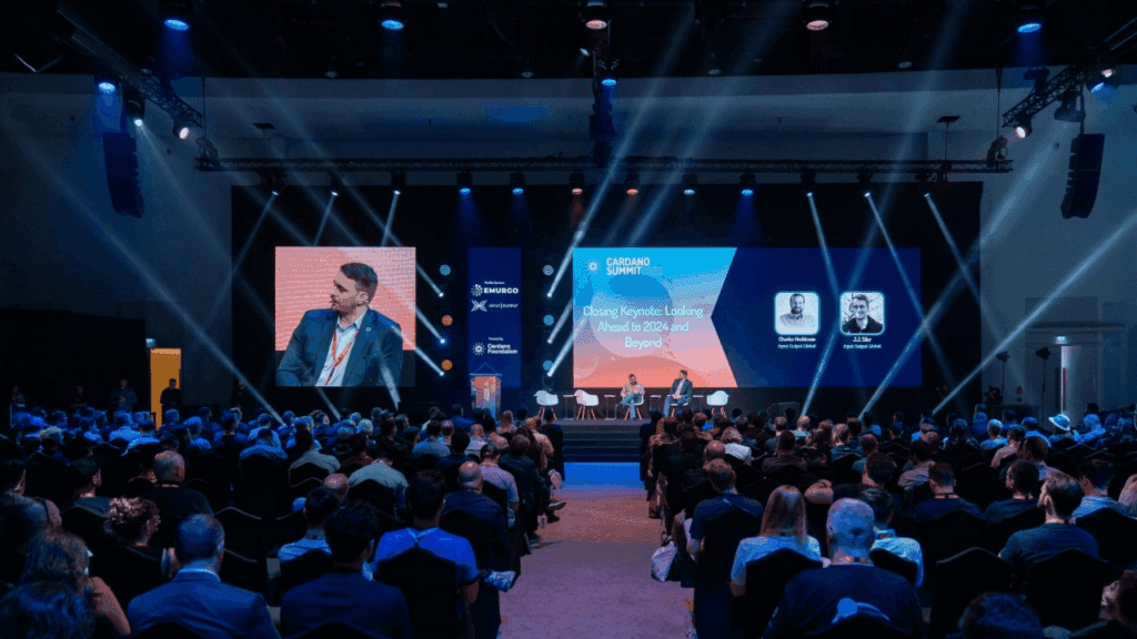 A large audience watches a keynote speaker on stage at a conference, with bright lights and a large screen displaying event information behind the speakers—expertly coordinated by the conference organiser.