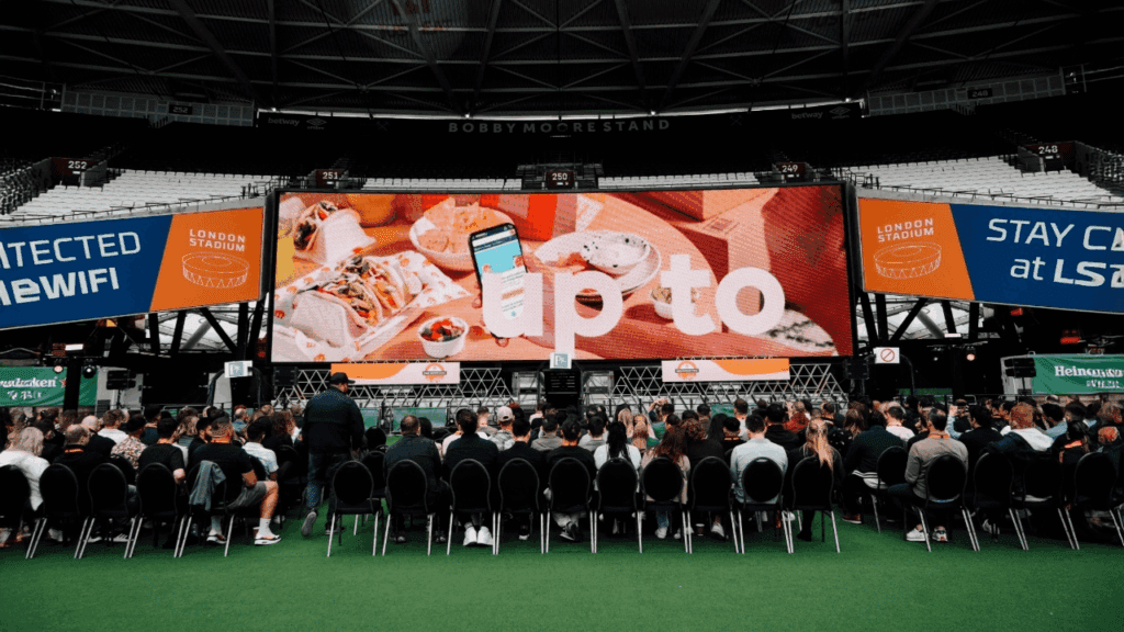 A large audience watches a keynote speaker on stage at a conference, with bright lights and a large screen displaying event information behind the speakers—expertly coordinated by the conference organiser.