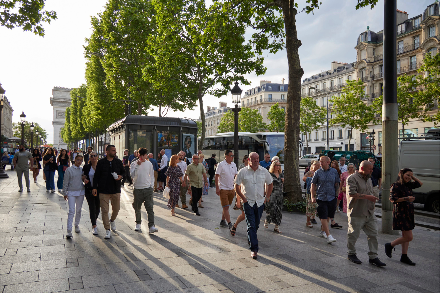 Restorative Experiences Blog. People walk along a sunny, tree-lined street in Paris near the Arc de Triomphe, enjoying restorative experiences amid lively buildings, vehicles, and a newsstand in the bustling scene.