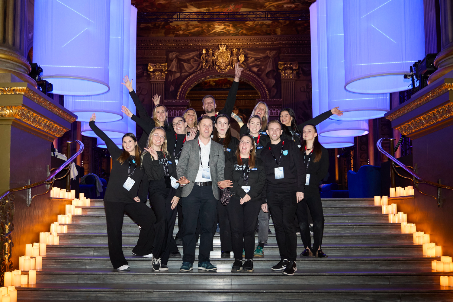 A group of people pose and smile on a grand staircase lined with candles at a corporate event agency gathering, in an ornate hall with golden decorations and large illuminated pillars. Most are wearing black outfits and conference badges.