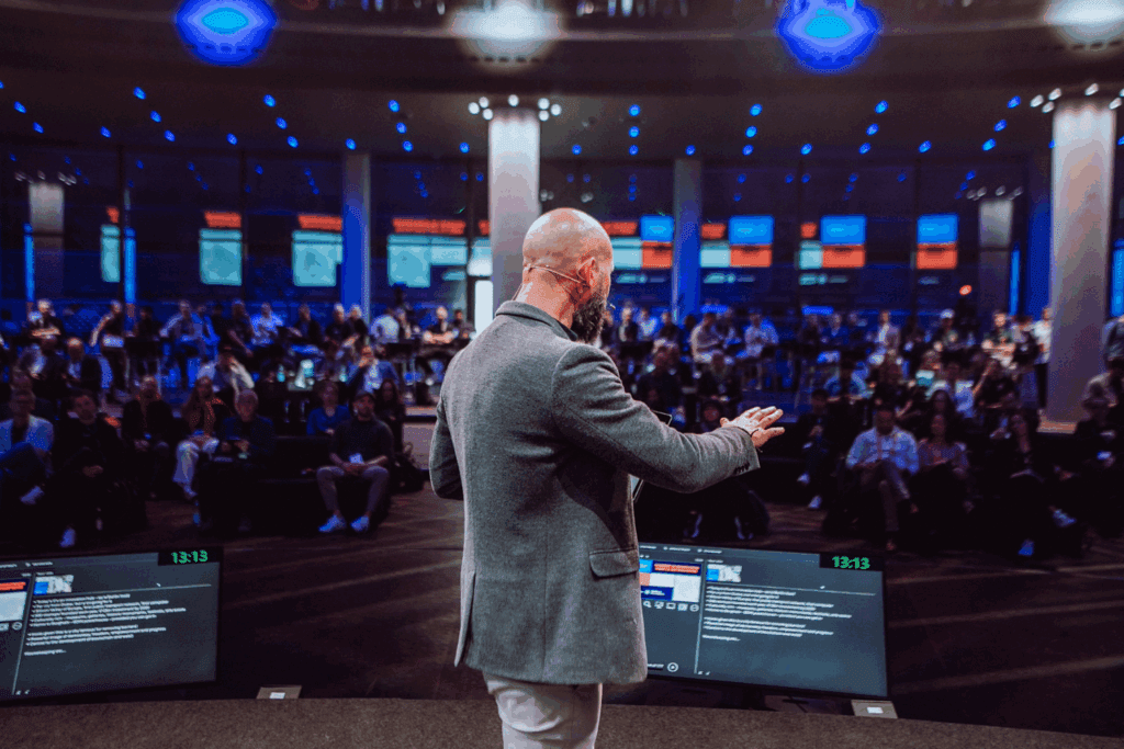 A man with a shaved head and beard, wearing a gray blazer, speaks on stage at the Cardano Summit to an audience in a large, modern conference hall with bright lights and multiple screens displaying information.