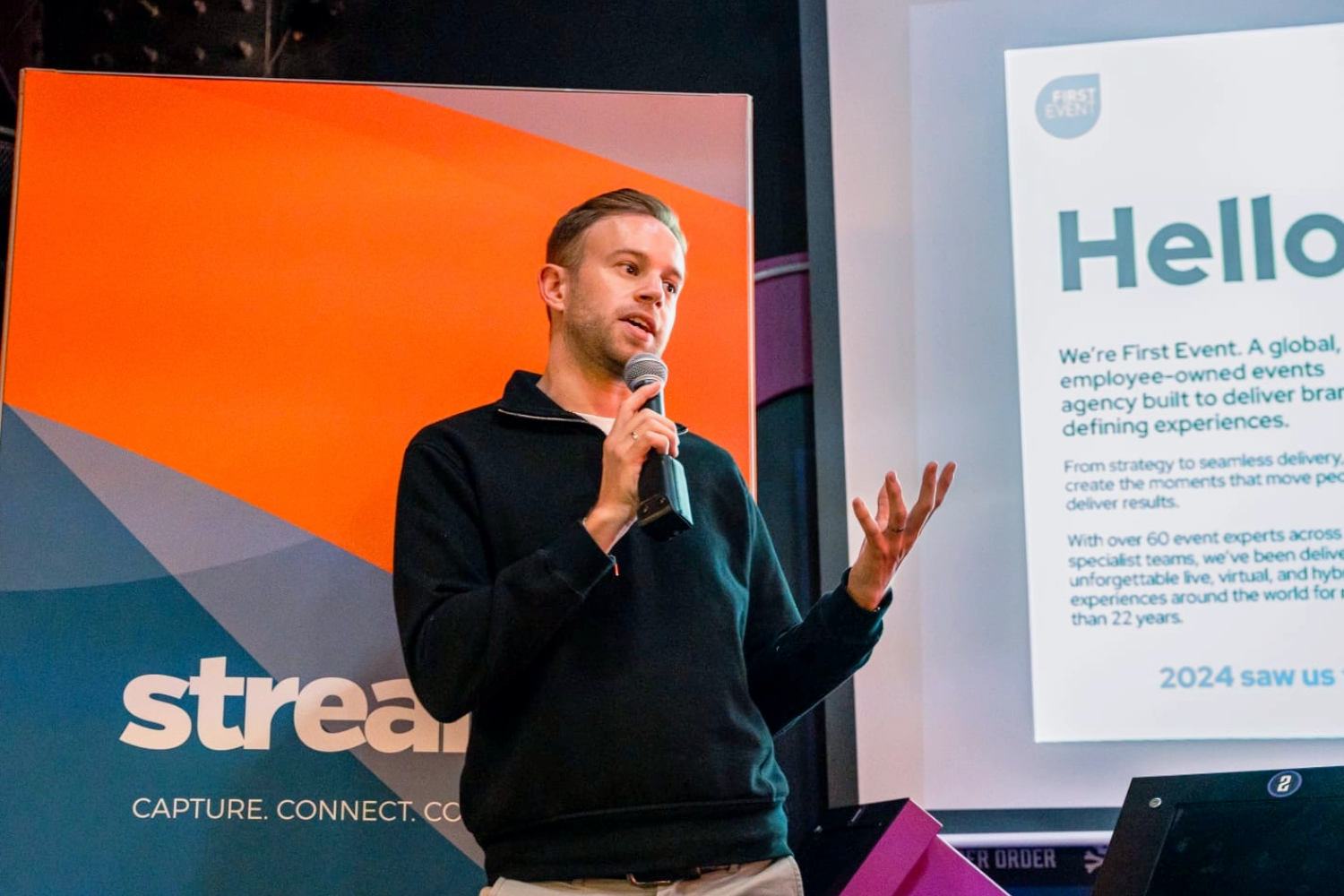 A man holding a microphone speaks on stage next to a screen displaying a presentation and a sign that reads stream. Capture. Connect. The screen behind him shows the word Hello.