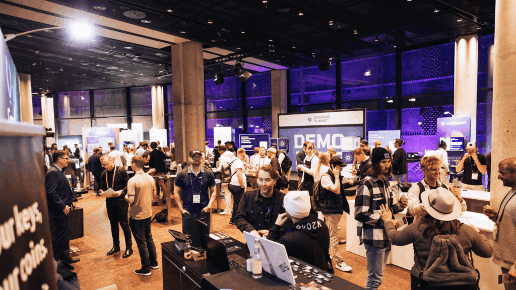 A large group of people interact and network at an indoor tech conference or expo, such as Cardano Summit, with various booths, demo displays, and informational banners visible under bright, modern lighting.