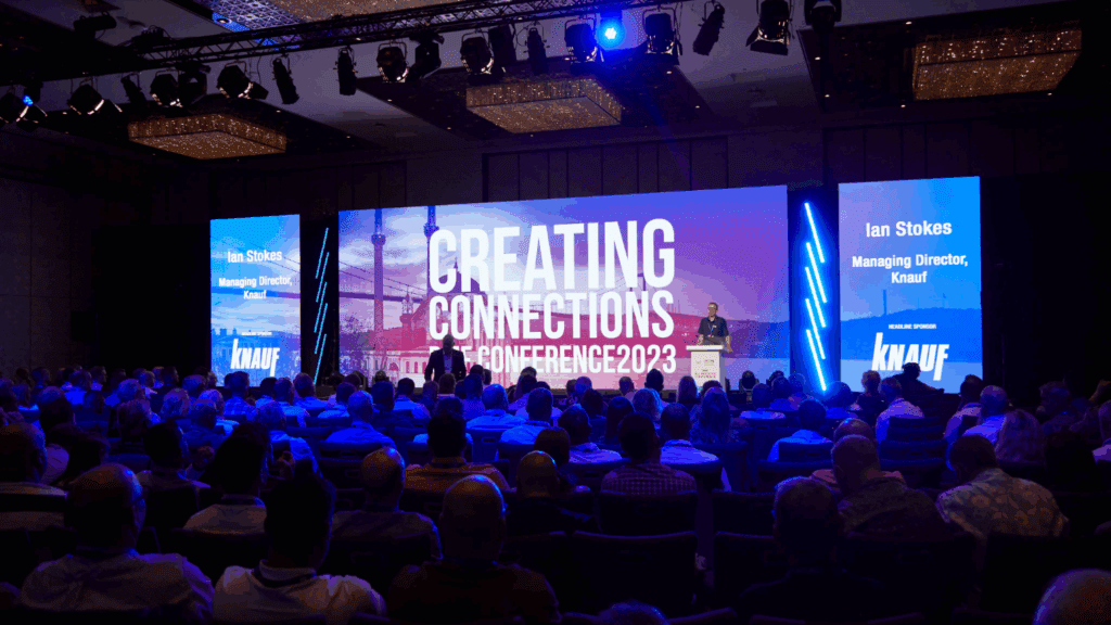A sustainable conference hall with a large audience faces a stage and screen displaying “Creating Connections THECONFERENCE2023.” Two side screens show “Ian Stokes, Managing Director, Knauf,” while blue and purple lighting sets the scene.