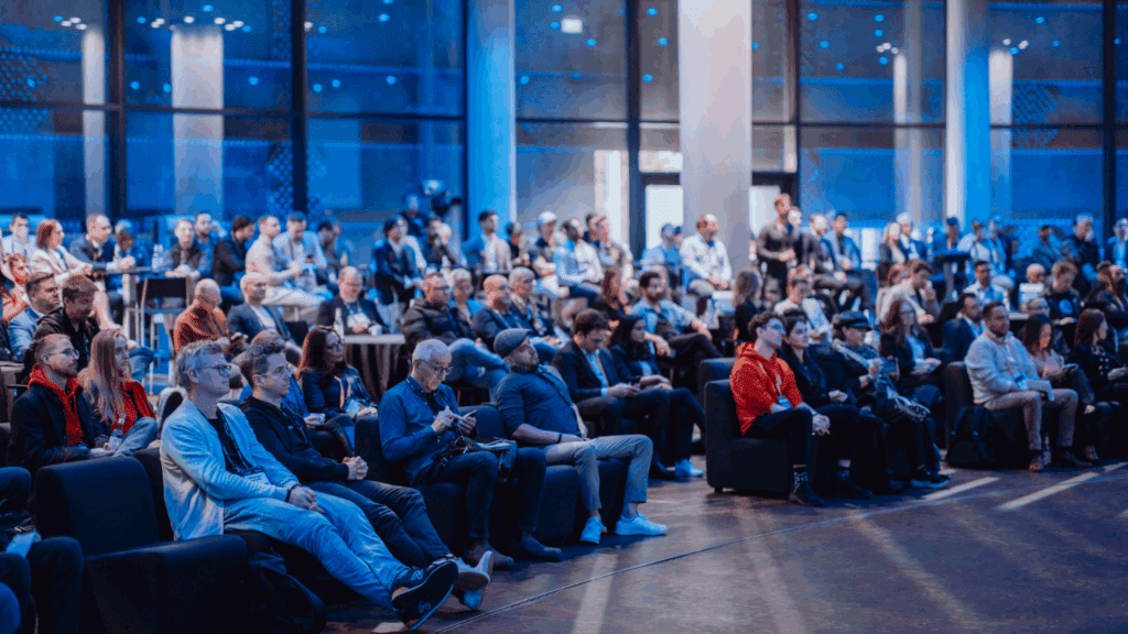 A large group of people seated and attentively watching an event or presentation at the Cardano Summit in a spacious, modern venue with blue lighting and tall windows.