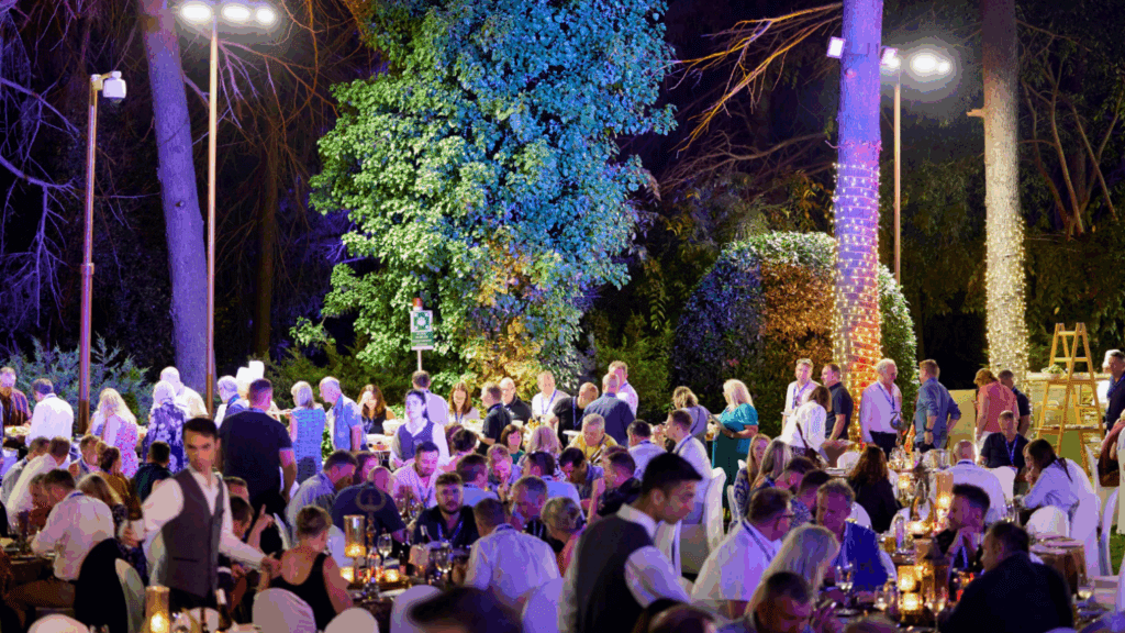 A large group of people gather outdoors at night for a festive, sustainable conference, seated at tables with candles and lights, surrounded by trees decorated with string lights.