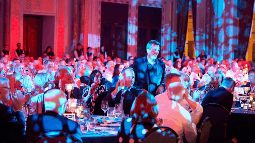 A man stands smiling and is being applauded by a large audience seated at round tables in a ballroom, lit with colorful, blue and red stage lights, creating a festive atmosphere at the sustainable conference.
