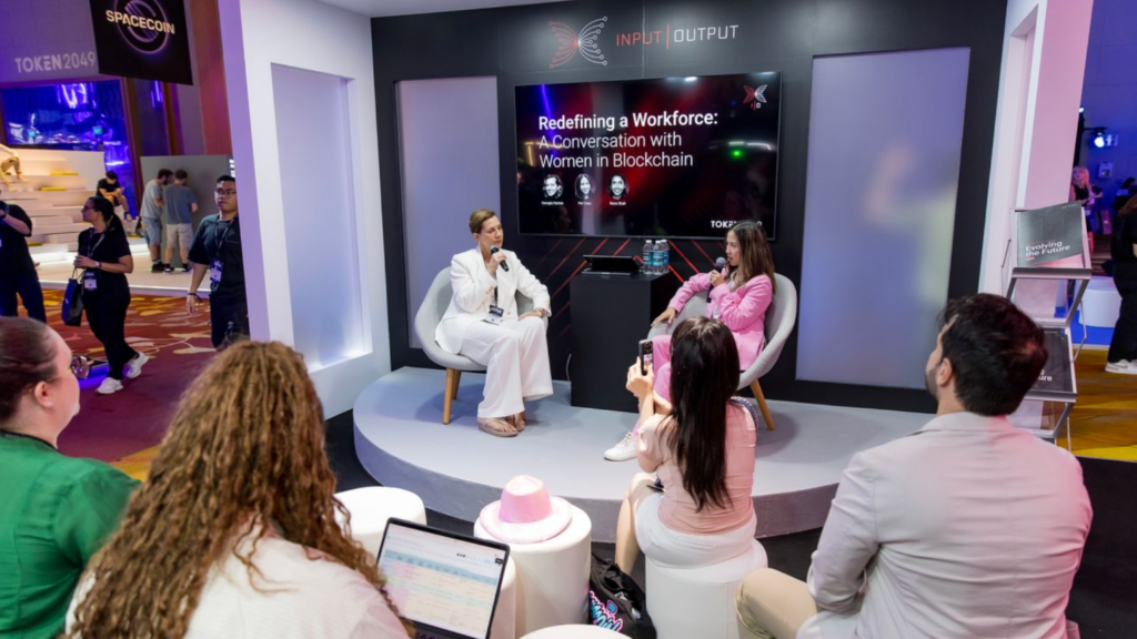 Two women sit on stage at an Expo Stand Build, having a discussion titled “Redefining a Workforce: A Conversation with Women in Blockchain,” while an audience listens and takes notes at the conference event.