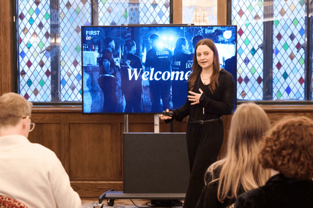 A woman stands and speaks in front of a screen displaying the word Welcome, addressing an audience attending the Unlocking Experiences webinar, with stained glass windows framing the room.