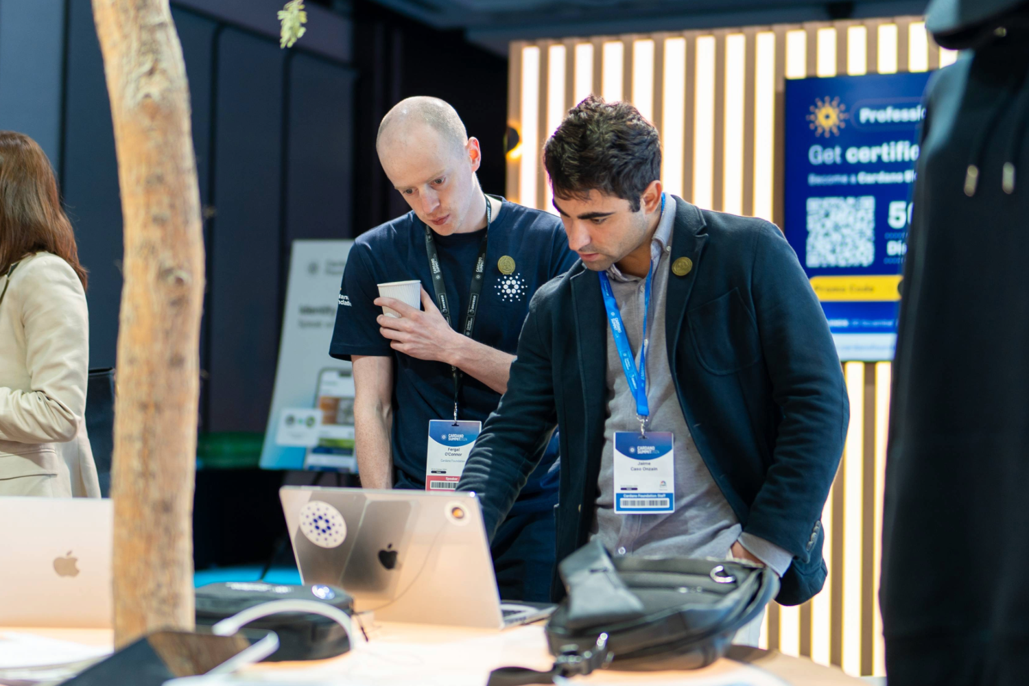 Two men at a tech event stand at a table with laptops. One checks his phone as the other points at a laptop screen featuring webinars on-demand. Both wear event badges and appear focused. A promotional sign is visible in the background.