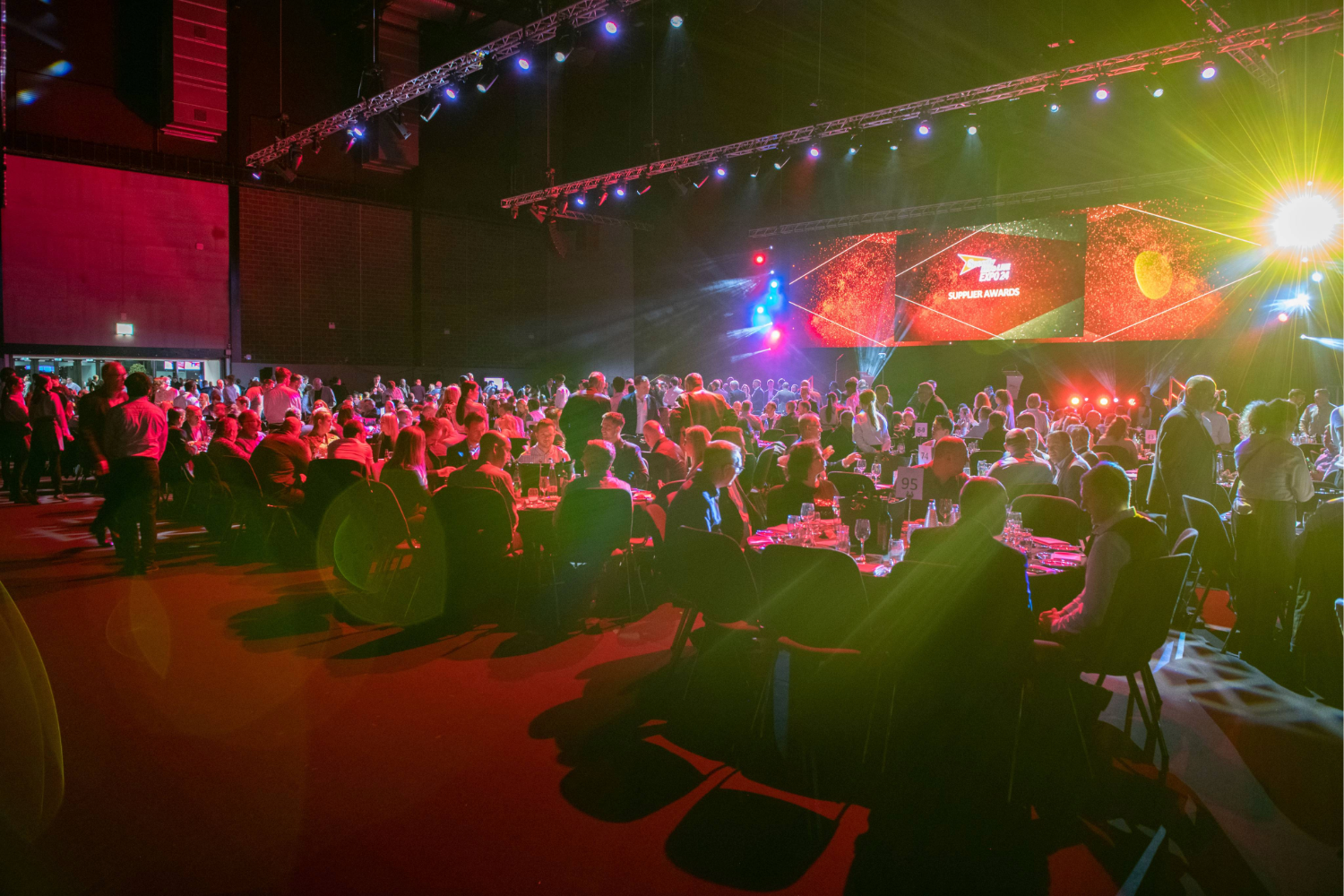 A large crowd sits at round tables in a dimly lit event hall, facing a brightly lit stage with colorful lights and a screen displaying graphics at what appears to be an awards ceremony or gala.