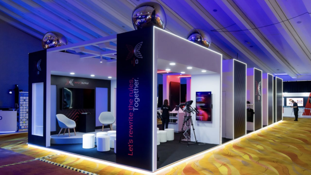 A modern Expo Stand Build featuring illuminated edges, white stools, tables, and display screens under a high ceiling. The booth showcases the slogan, Let's rewrite the rules. Together! in prominent lettering.