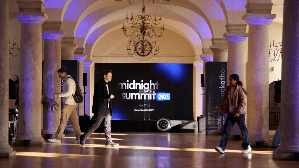 People walk through an elegant hall with chandeliers and columns, immersed in a striking brand experience. A large screen displays midnight summit 2025 and event details, while blue and purple lighting sets a modern, professional mood.