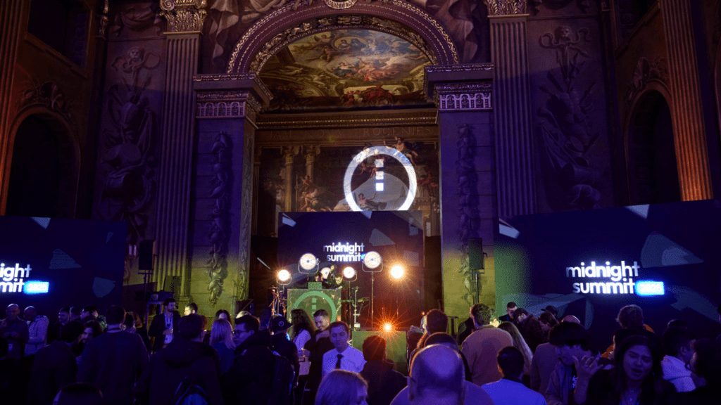 A crowd gathers in a grand, ornately decorated hall with high ceilings and murals, attending an immersive brand experience event with stage lighting and screens displaying “midnight summit 2022.”.
