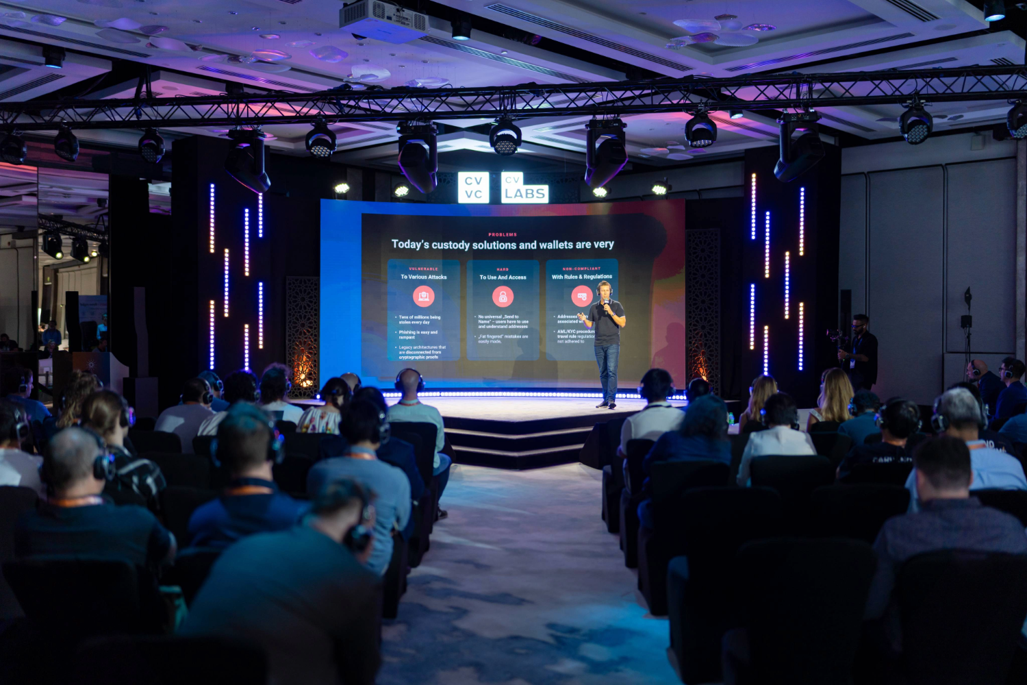 A speaker stands on stage presenting creative event services to an attentive audience in a conference room, with a large screen behind displaying information about custody solutions and wallets.