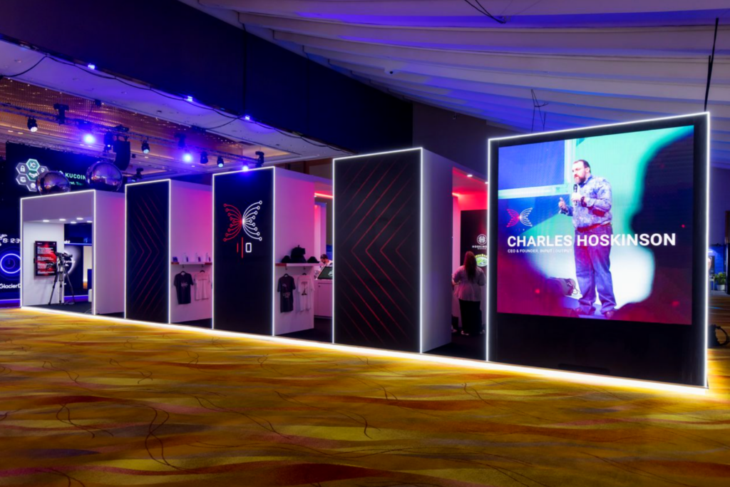 A modern Expo Stand Build featuring illuminated edges, white stools, tables, and display screens under a high ceiling. The booth showcases the slogan, Let's rewrite the rules. Together! in prominent lettering.