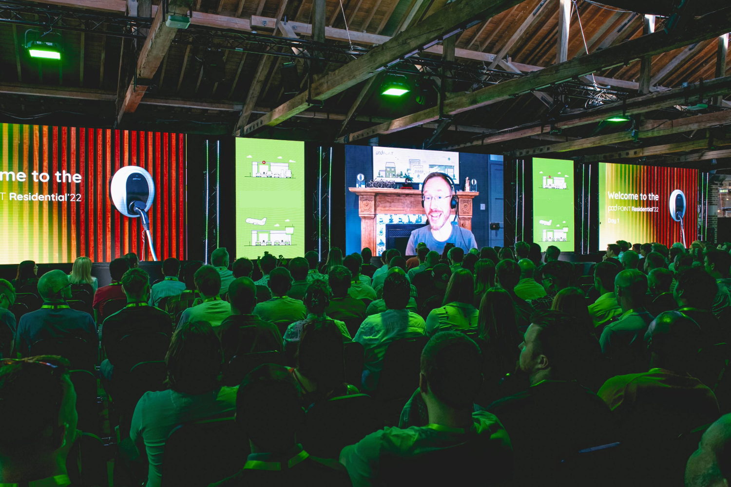 A large audience sits in a darkened venue, facing a stage with multiple screens showing a smiling person speaking virtually. Green lighting and event marketing visuals illuminate the crowd, while beams shimmer on the wooden ceiling above.