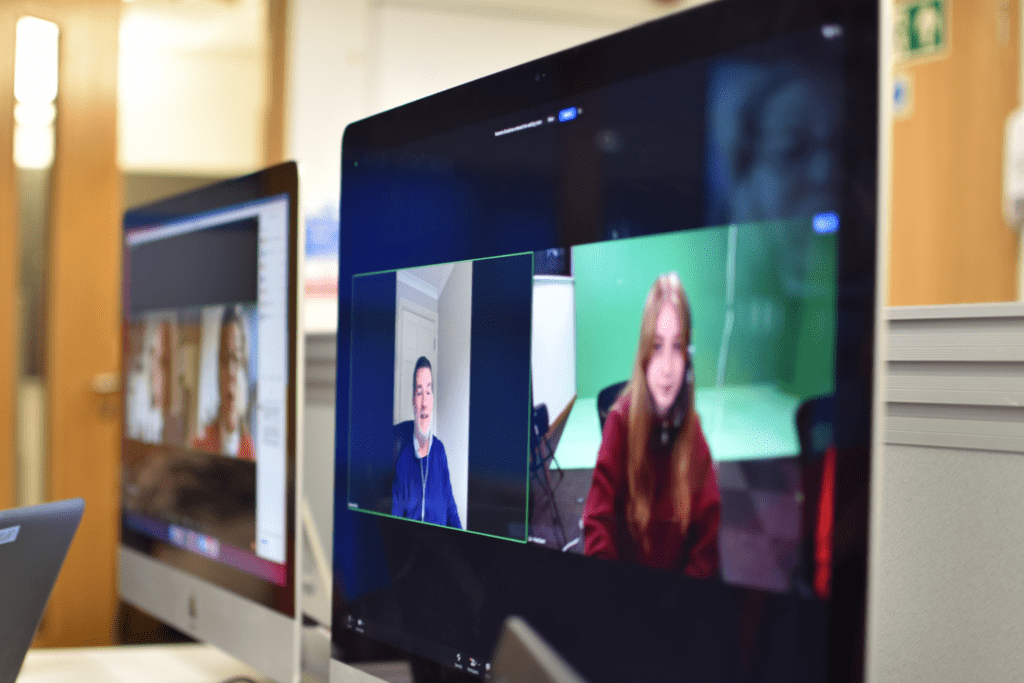 A computer monitor shows a video call with two people, one in a blue shirt and one in a red jacket, whilst other screens display virtual events in the background of an office setting.