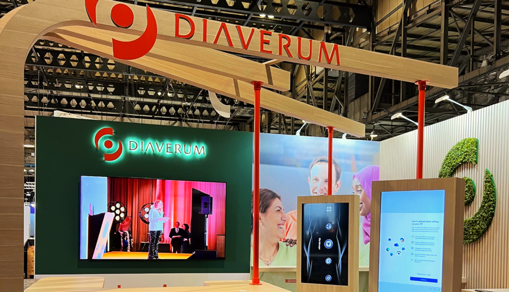 A modern exhibition stand for Diaverum features a large screen displaying a speaker, a smaller digital touchscreen, brand logos, and images of smiling people, highlighting shared experiences under a geometric wooden and red frame overhead.