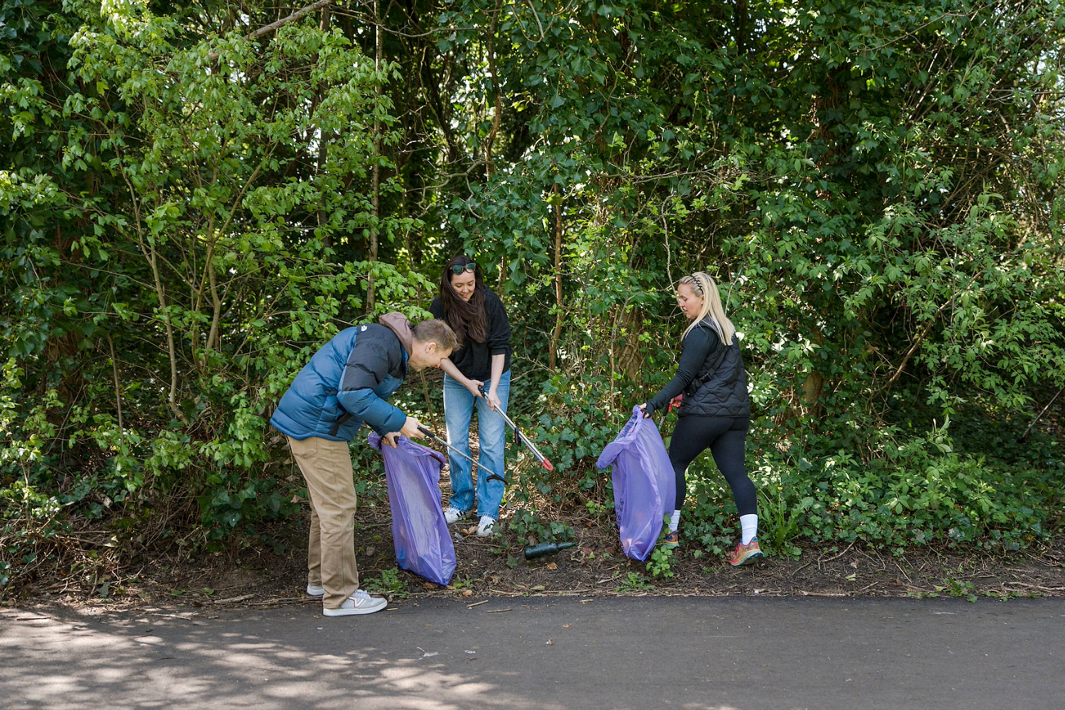 Three people collect litter along the edge of a wooded area, using litter pickers and holding large purple bags. Trees and bushes surround them, and they stand on a path next to the greenery.
