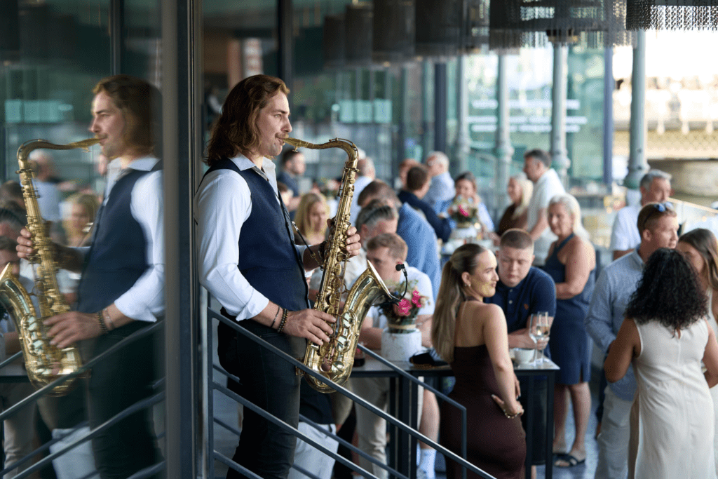 A saxophonist plays at an upmarket indoor-outdoor event, reflected in glass beside him, as well-dressed guests enjoy drinks and engage in shared experiences, mingling and conversing throughout the evening.
