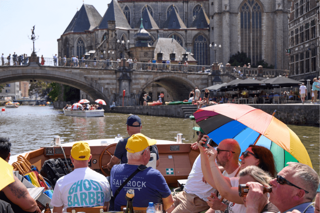 A group of people share experiences as they ride in a small boat on a canal, some taking photos and holding a rainbow umbrella. A stone bridge and large gothic church are visible in the background, with people relaxing by the water.